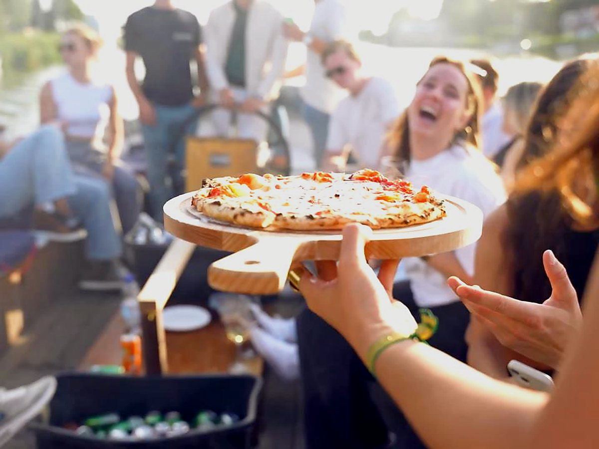 Pas gebakken pizza op een houten schep aan boord, met gasten op de achtergrond